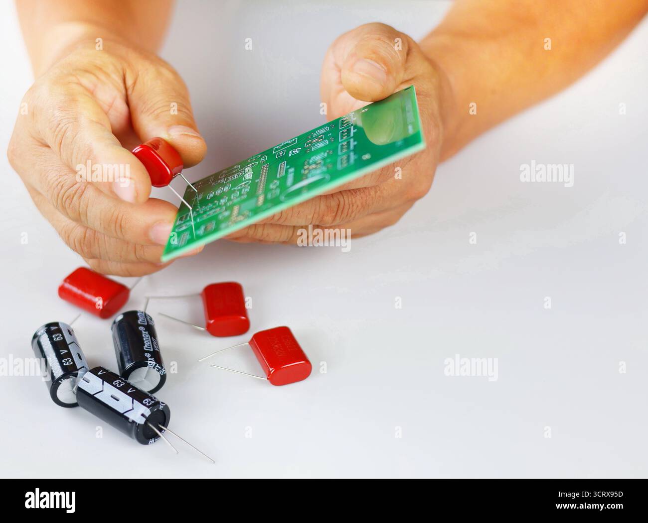 Die Abbildung zeigt ein rotes elektronisches Bauteil, wahrscheinlich ein Kondensator oder Widerstand, das zur Montage auf einer grünen Leiterplatte (PCB) in der Hand gehalten wird. Stockfoto