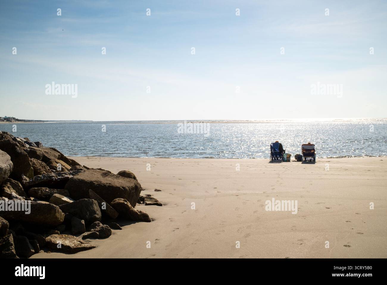 Liegestühle am Isle of Palms Beach South Carolina // SULLIVANS ISLAND, South Carolina — zwei Personen entspannen sich in Liegestühlen am Sullivans Island Beach, mit dem Wasser von Breach Inlet im Vordergrund. Die ferne Küste der Isle of Palms ist über den Einlass sichtbar. Sowohl Sullivans Island als auch Isle of Palms sind Barriereinseln in der Nähe von Charleston, South Carolina, die für ihre Strände und Freizeitaktivitäten bekannt sind. Stockfoto