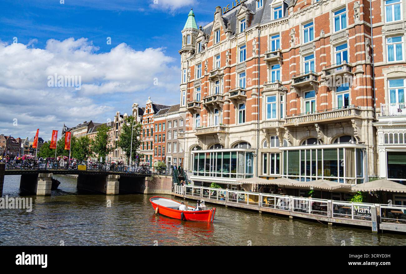 Rotes Boot auf einem Kanal in Richtung einer Brücke, auf der Menschen laufen, Fahrräder parken, Wohnhäuser und Cafés mit Sonnenschirmen in Amsterdam, Niederlande, Stockfoto