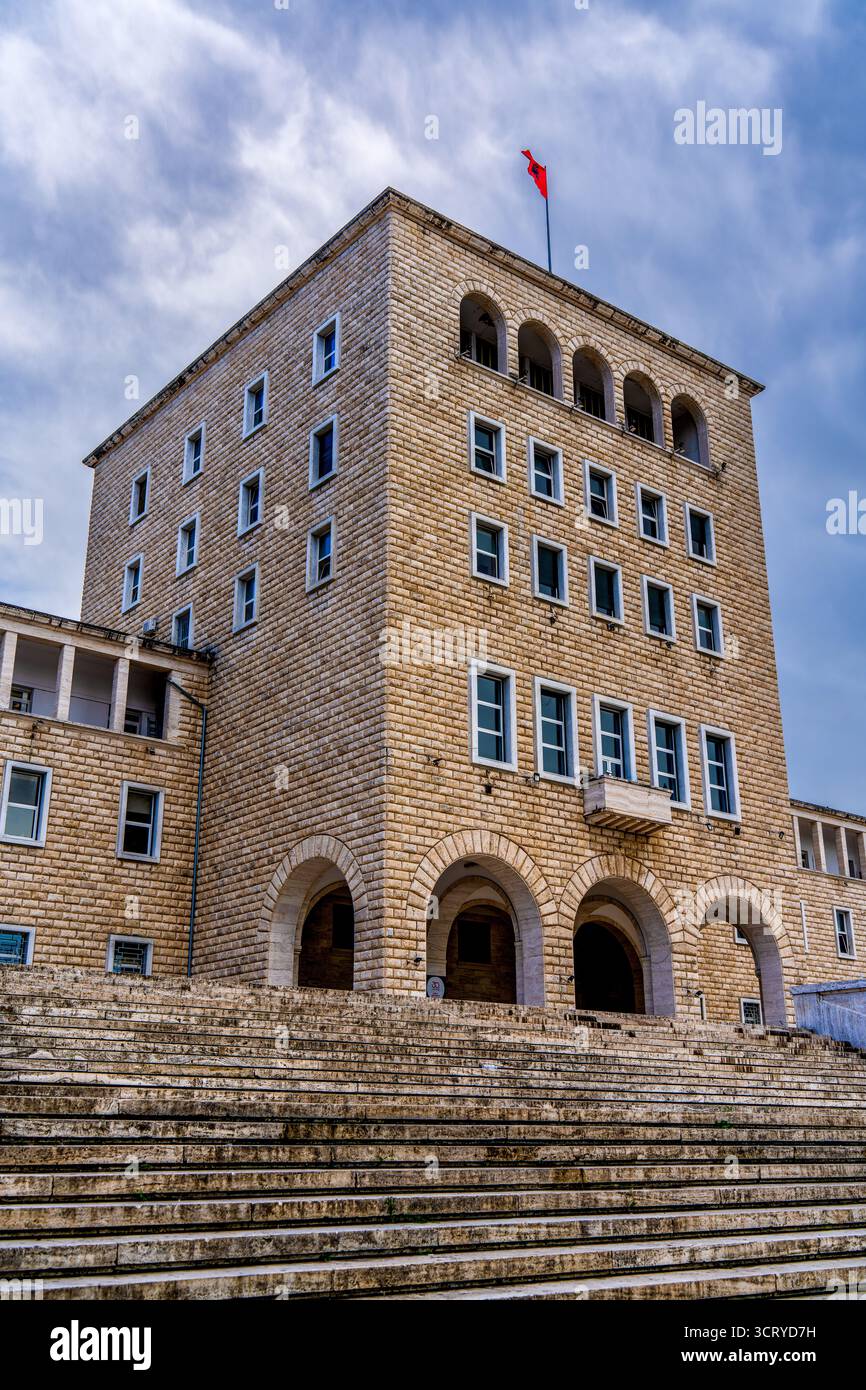 Historisches Steingebäude der Universität von Tirana mit bogenförmigem Eingang, Fenstern und albanischer Flagge unter bewölktem Himmel Stockfoto