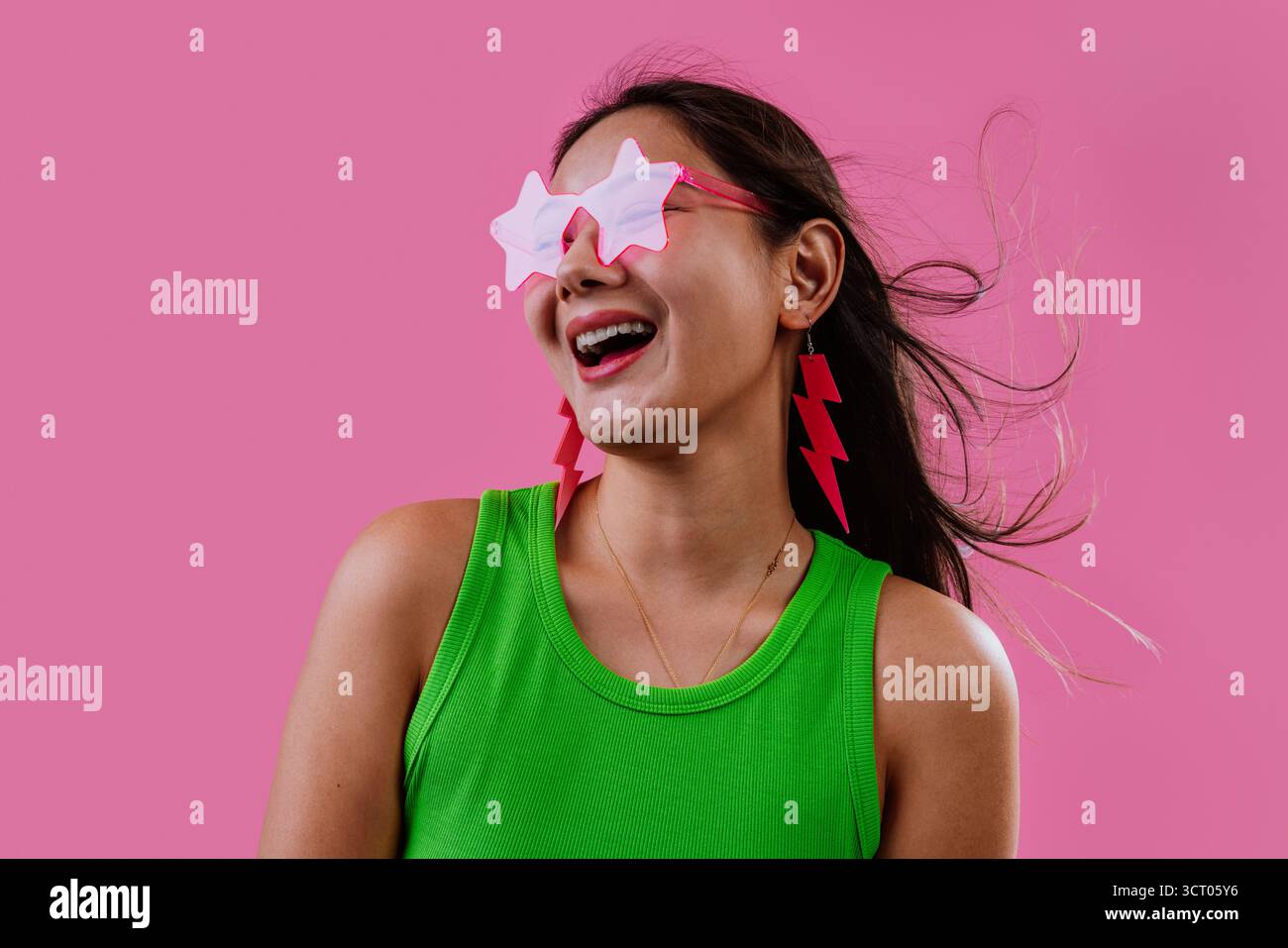 Junge Frau, die fröhlich lächelt in rosa Sonnenbrille und blitzschnellen Ohrringen, Haare wehen vor einem leuchtenden rosa Hintergrund, verspielt, trendig, karg Stockfoto