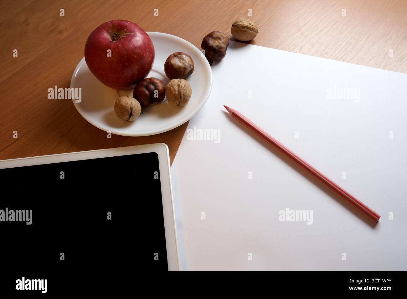 Ein roter Apfel und Nüsse sitzen auf einem Teller neben leerem Papier und einem farbigen Bleistift. Stockfoto
