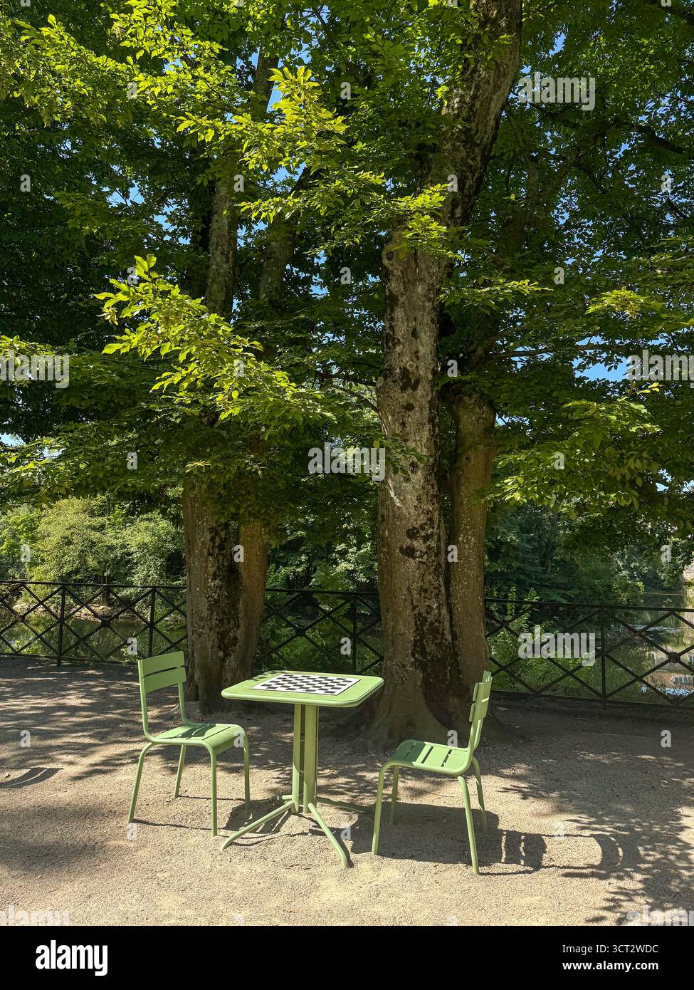 Zwei Stühle und ein grüner Tisch mit einem Schachbrett unter grünen Bäumen in einem öffentlichen Park, mit Sonnenlicht, das durchströmt, und Doubs River im Hintergrund - Smartphone-aufgenommenes Stockfoto