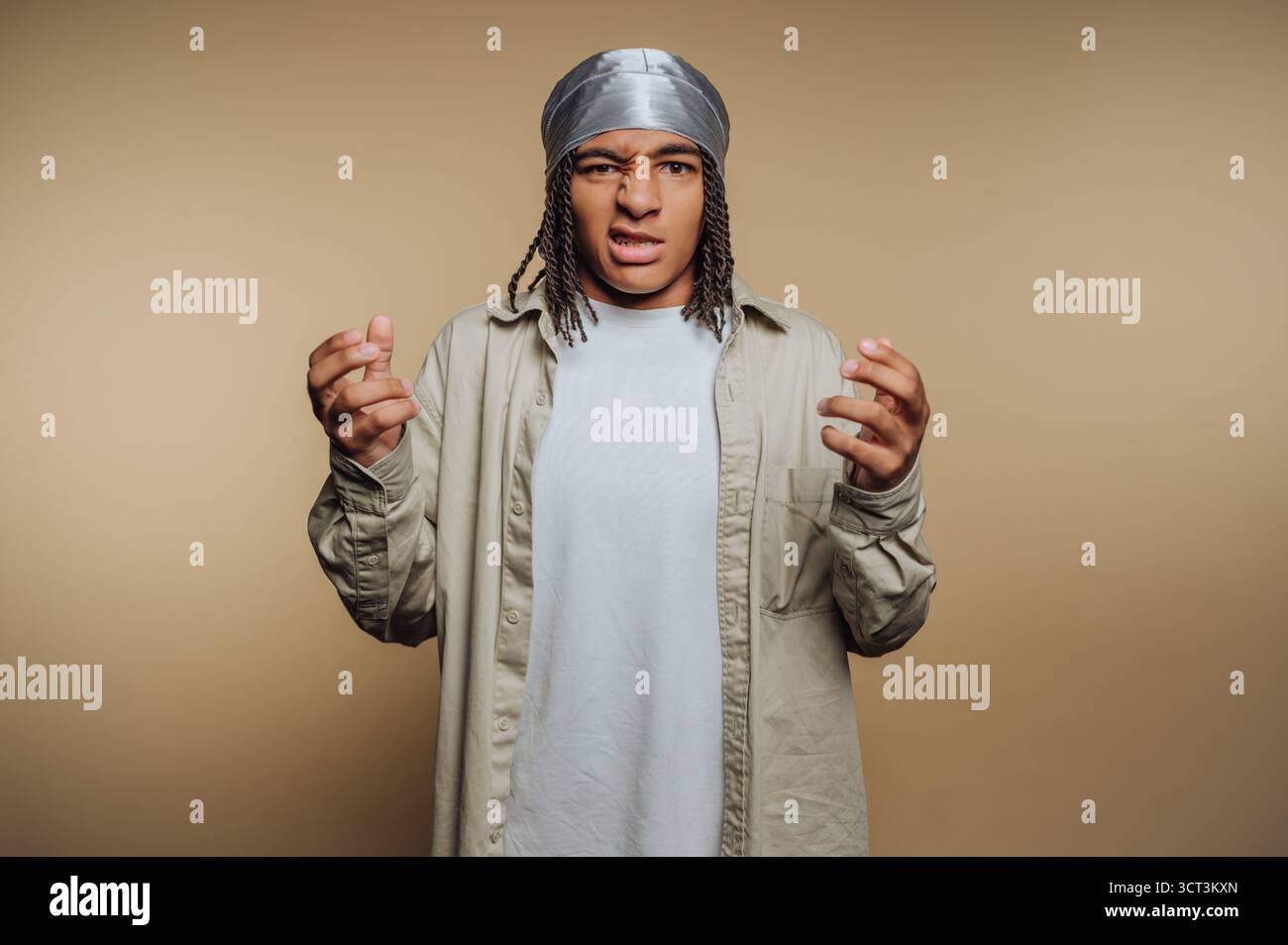 Ein junger Mensch steht vor einem schlichten Hintergrund und schaut frustriert aus. Sie tragen ein lässiges Outfit und eine silberne Kopfpackung, die mit ihrem Ha Gesten gibt Stockfoto