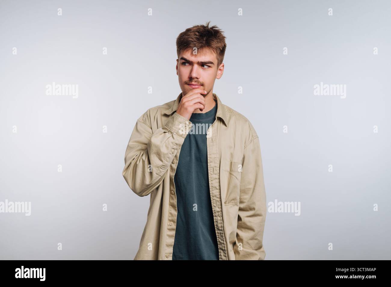Ein junger Mann steht in einem hell beleuchteten Studio und trägt ein lässiges Outfit. Er hat einen nachdenklichen Ausdruck, als er sein Kinn berührt, verloren in der Nachdenklichkeit abo Stockfoto