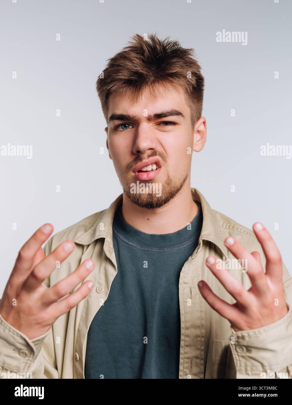 Ein junger Mann steht in einem hellen Atelier und zeigt einen frustrierten Ausdruck. Seine Hände sind hochgehoben, als ob er Antworten sucht, und er trägt ein lässiges Outfit Stockfoto