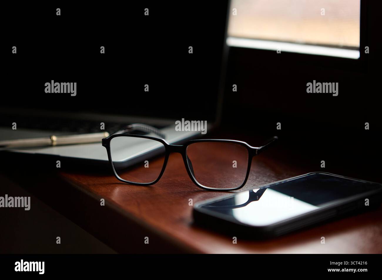 Die elegante schwarze Brille liegt auf einer polierten Schreibtischoberfläche mit Smartphone und Laptop im Hintergrund auf und sorgt so für ein schlankes, minimales Bürodesign. Stockfoto