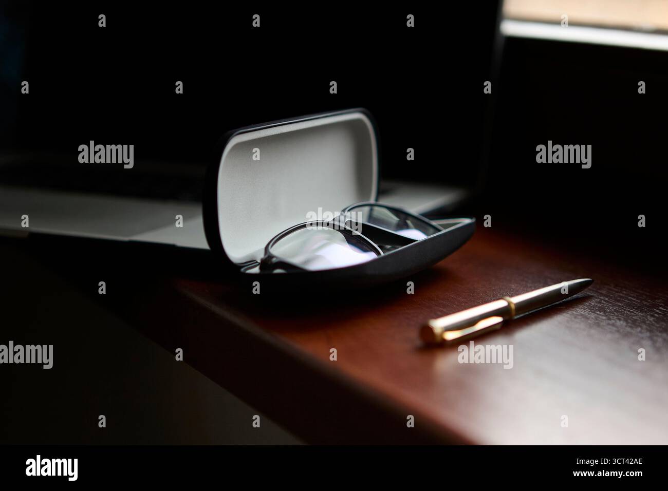 Offenes Brillenetui mit Brille im Inneren auf poliertem Holzschreibtisch neben Stift und verschwommenem Computer im Hintergrund. Sauberes Produkt, Stillleben. Stockfoto