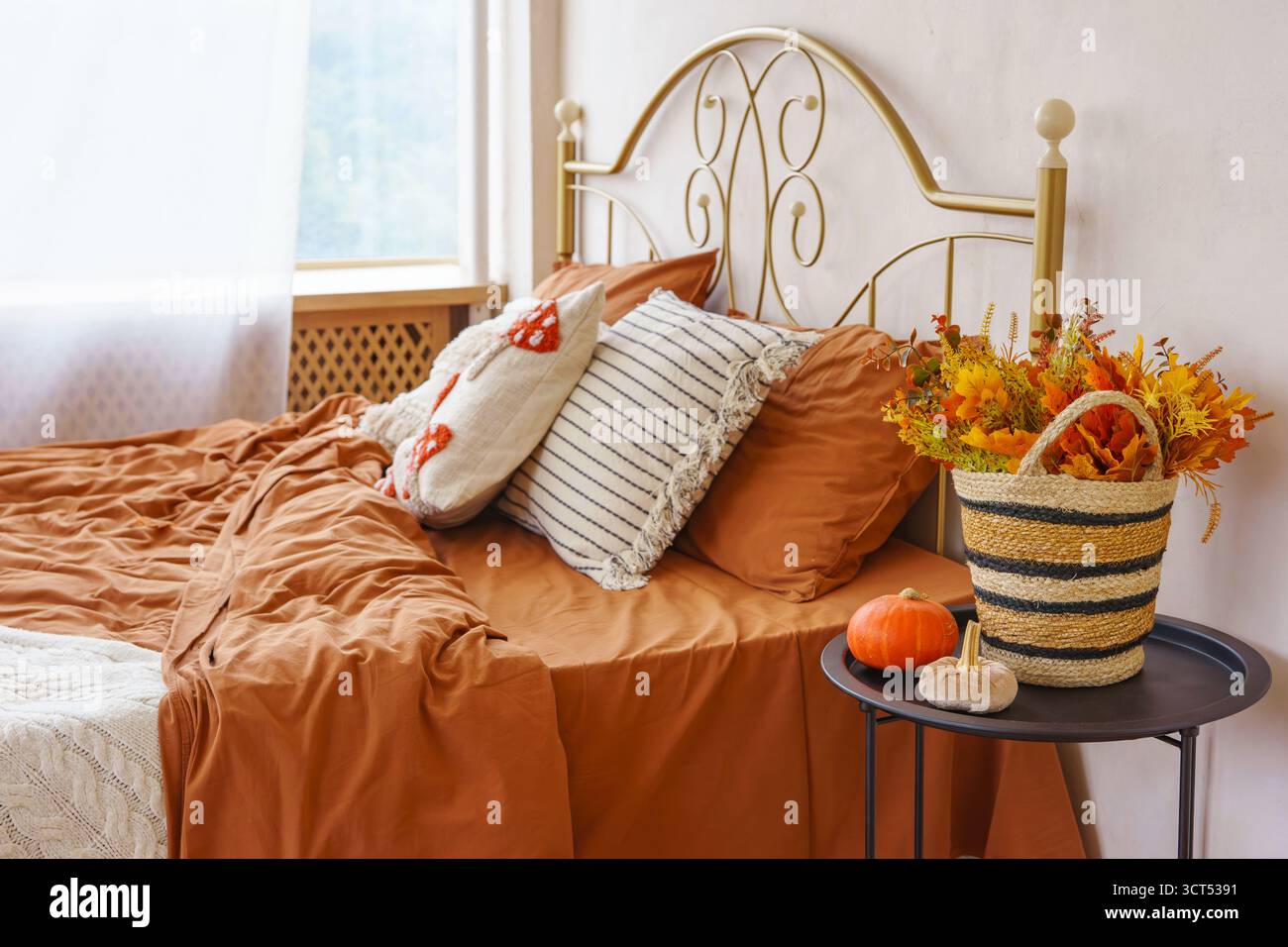 Schlafzimmer mit warmbraunem Bettzeug, Kissen, Korb mit Herbstblättern und Kürbissen, die eine gemütliche, saisonale Inneneinrichtung schaffen. Geschmiedetes Bettkopfteil, Braue Stockfoto