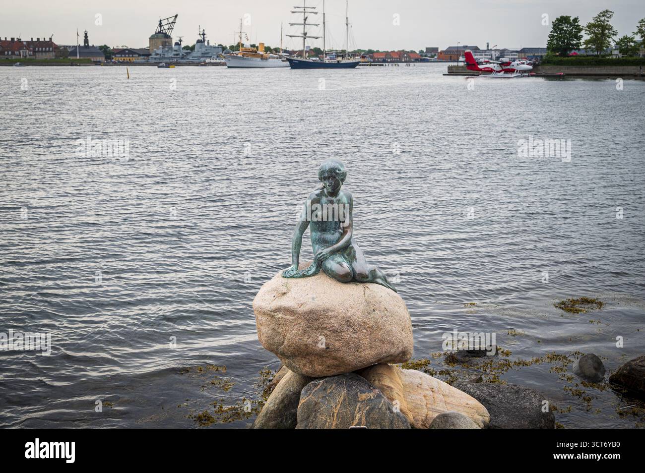 Bronzene Meerjungfrau auf einem Felsen mit Schiffen und Hafen im Hintergrund, Kopenhagen, Dänemark Stockfoto