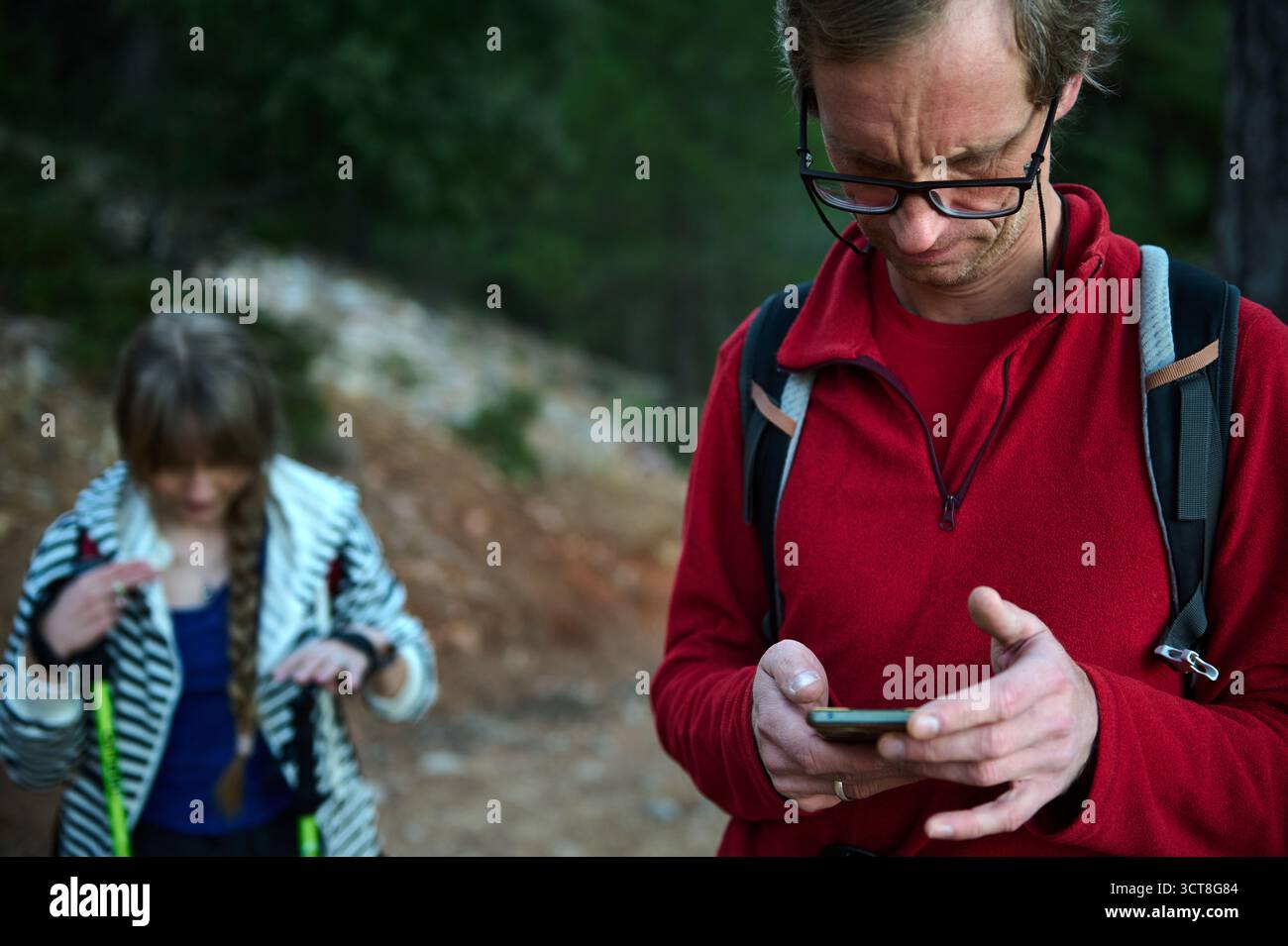 Mann in rotem Fleece mit Brille überprüft sein Handy auf einem Waldweg, während eine Frau in gestreifter Jacke die Ausrüstung im Hintergrund, Rucksäcke, Wandern, Stockfoto