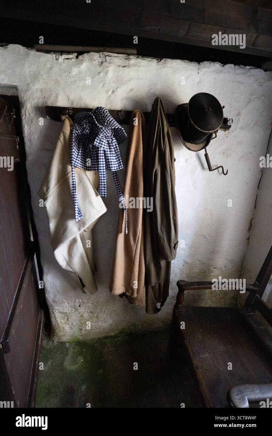 Historisches Cottage-Interieur mit alter Kleidung und Kopfhut, die an der weiß getünchten Wand hängen Stockfoto