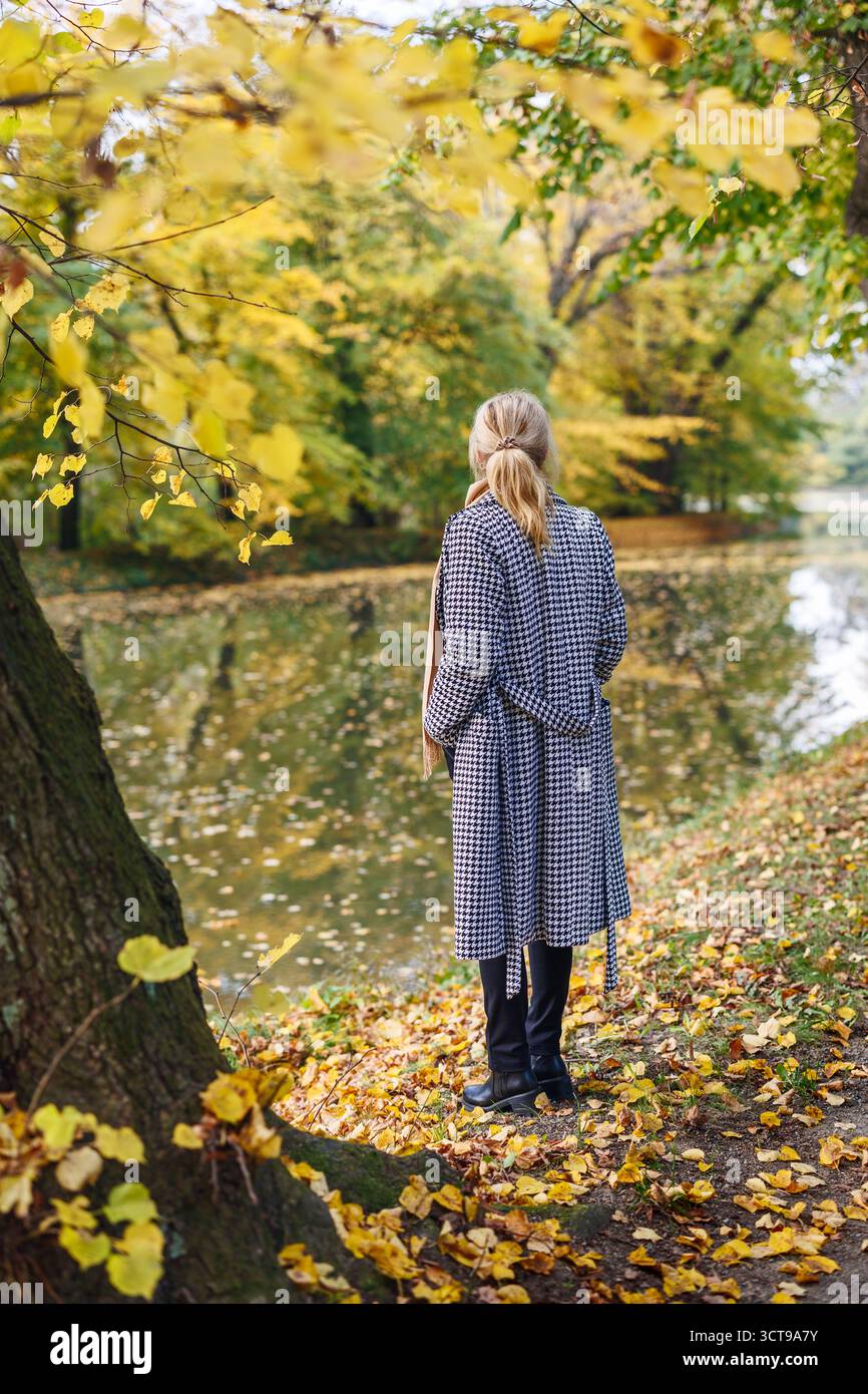 Eine nachdenkliche Frau im Hahnentrittmantel genießt Achtsamkeit und Einsamkeit im Herbstpark mit goldenen Blättern und friedlicher Natur Stockfoto
