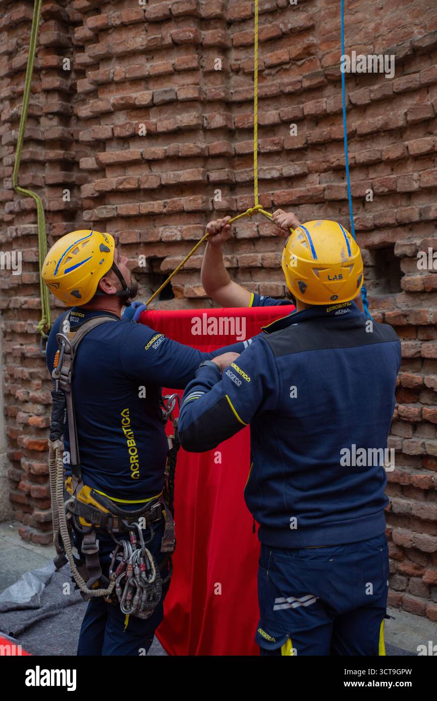 Seilzugangstechniker der Firma Acrobatica arbeiten zusammen, um eine große, leuchtend rote Leinwand an der Steinfassade des Santa M anzuheben und zu befestigen Stockfoto