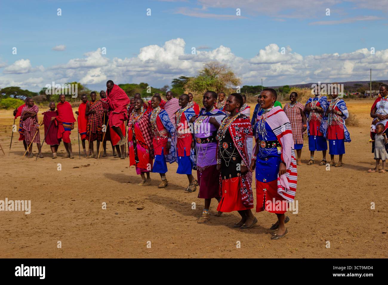 Maasai-Männer springen, während Frauen in einer Reihe laufen, die in farbenfroher traditioneller Kleidung verziert ist, während einer Zeremonie in ihrem kenianischen Dorf. Stockfoto