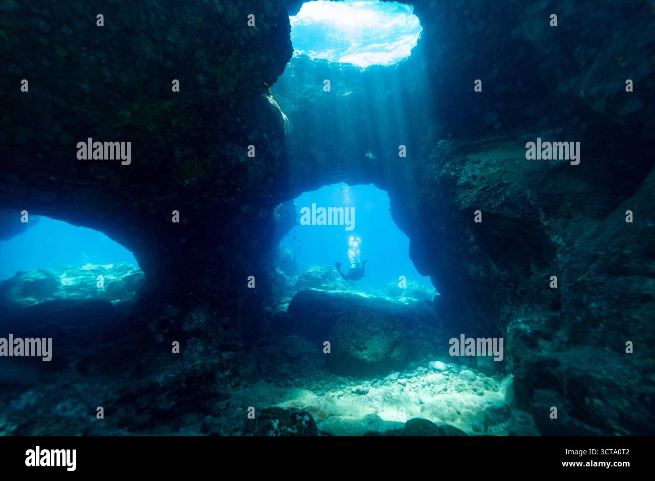 Eine Unterwasserszene zeigt einen Taucher, der eine Höhle erforscht, wobei das Sonnenlicht von oben hereinströmt und das türkisfarbene Wasser und die Felsformationen beleuchtet. Stockfoto