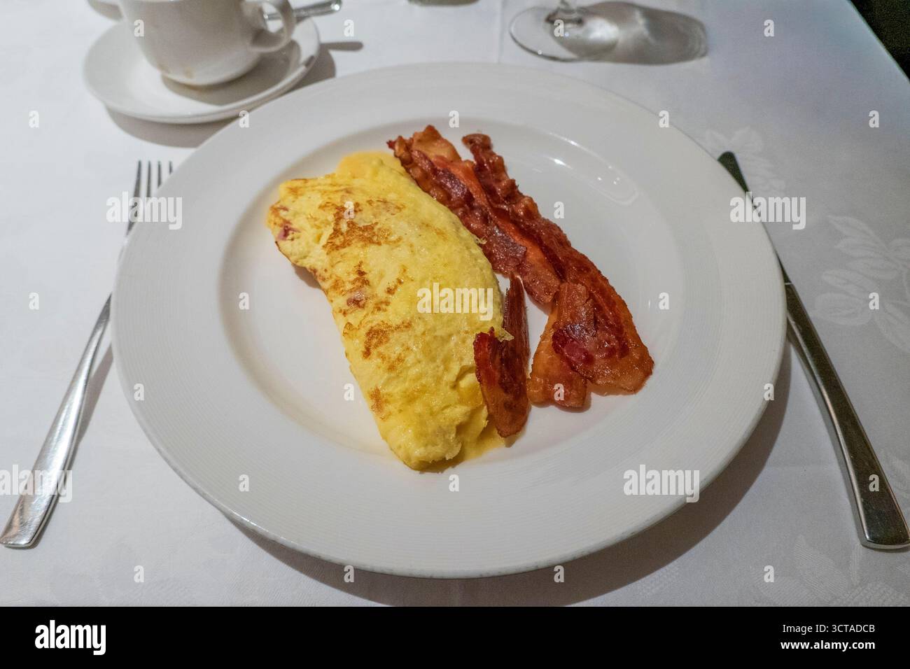Frühstücksteller mit flauschigem Omelett und knusprigen Speckstreifen, elegant auf einer weißen Tischdecke mit Besteck und einer Kaffeetasse präsentiert Stockfoto