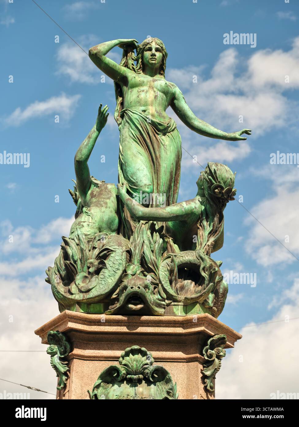 Bronzestatue, die eine Frau mit fließenden Haaren und ausdrucksstarker Pose darstellt, umgeben von mythologischen Figuren in einem klaren blauen Himmel Stockfoto