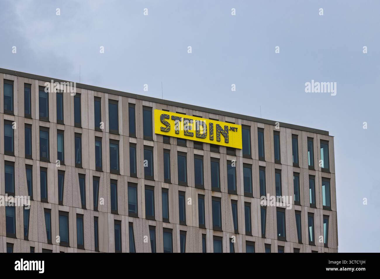 Die obere Ecke des modernen Stedin Bürogebäudes mit seiner markanten Fassade und dem leuchtend gelben Logo des Unternehmens vor grauem Himmel. Rotterdam Stockfoto