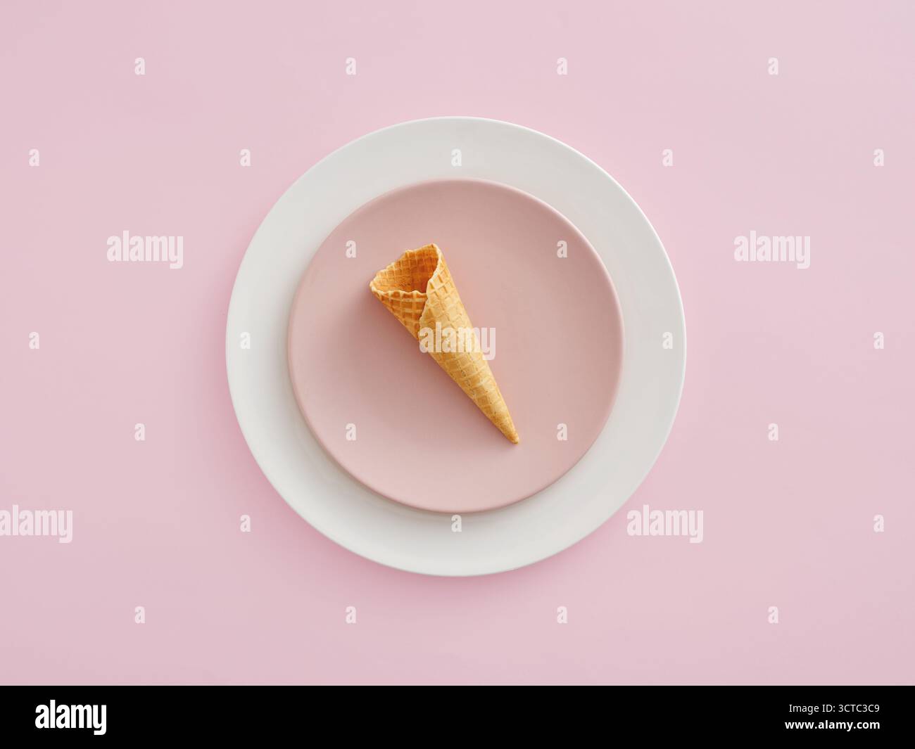 Leerer Waffelkegel auf einem rosafarbenen Teller, auf einem größeren weißen Teller mit pastellrosa Hintergrund. Minimales Dessertkonzept Stockfoto