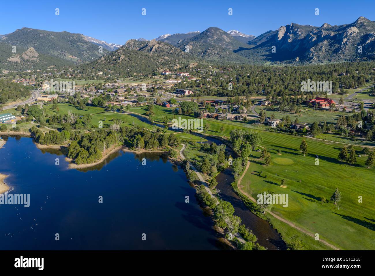 Aus der Vogelperspektive auf einen ruhigen See, der den Himmel spiegelt, umgeben von üppig grünen Golfplätzen, mit der Stadt eingebettet in die majestätischen, schneebedeckten Rocky Mountains, Estes Park, Colorado, USA. Stockfoto