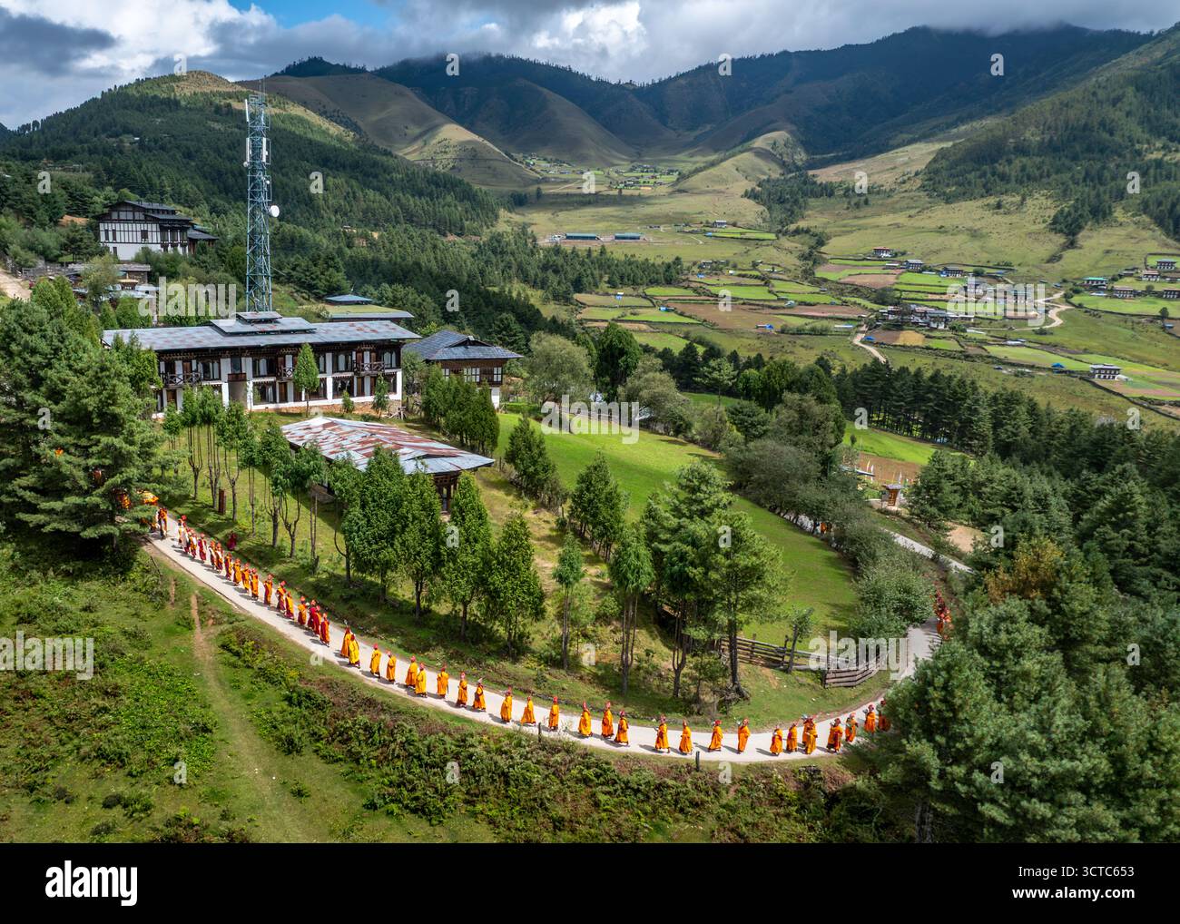 Aus der Vogelperspektive ziehen sich Mönche in Safrangewändern entlang eines Weges in der Nähe eines modernen Gebäudes vor dem Hintergrund der grünen Berge, Phobjikha, Wangdue Phodrang, B Stockfoto