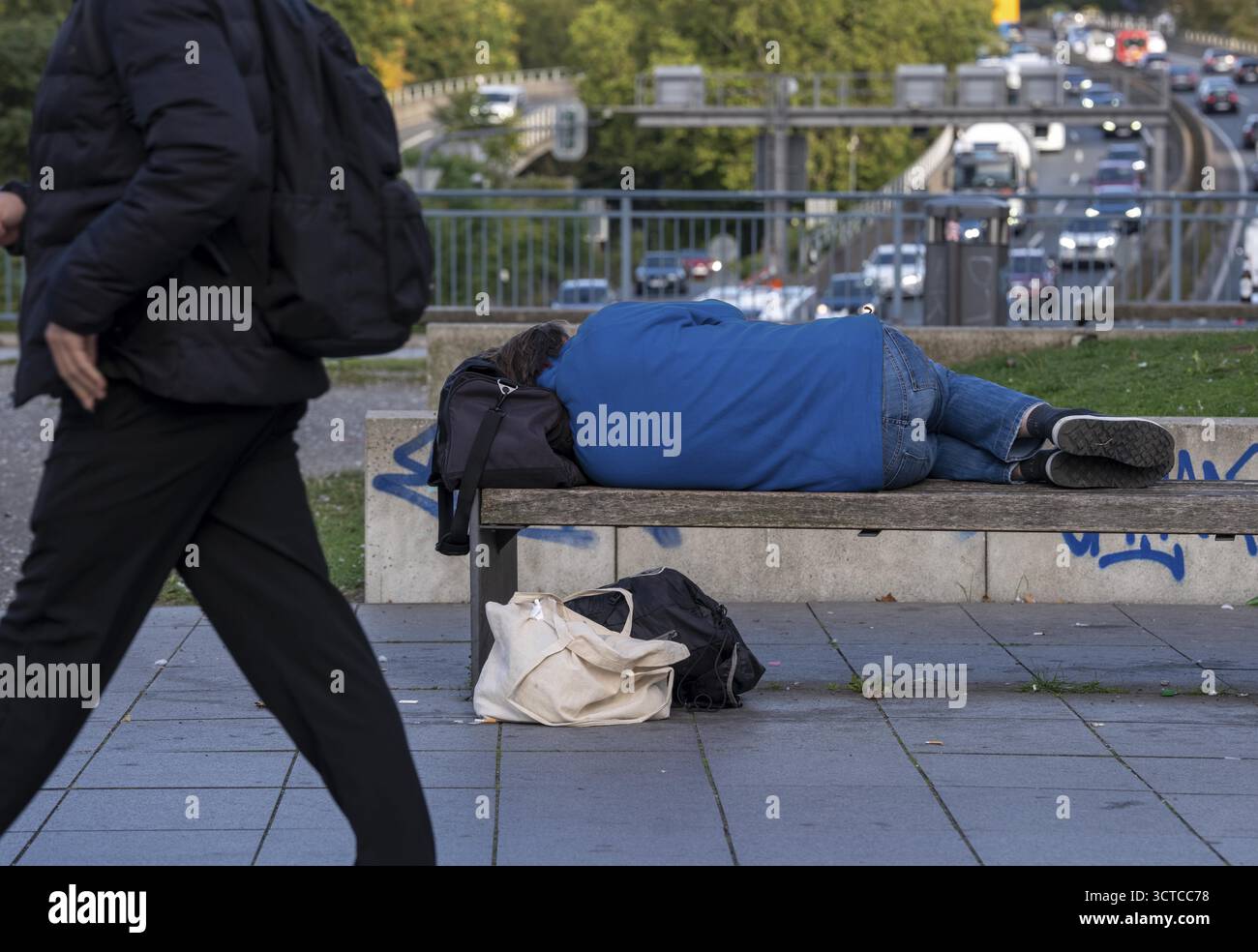 Mann schläft auf einer Bank, Reisetasche dient als Kissen, im Stadtzentrum spazieren Passanten vorbei Stockfoto
