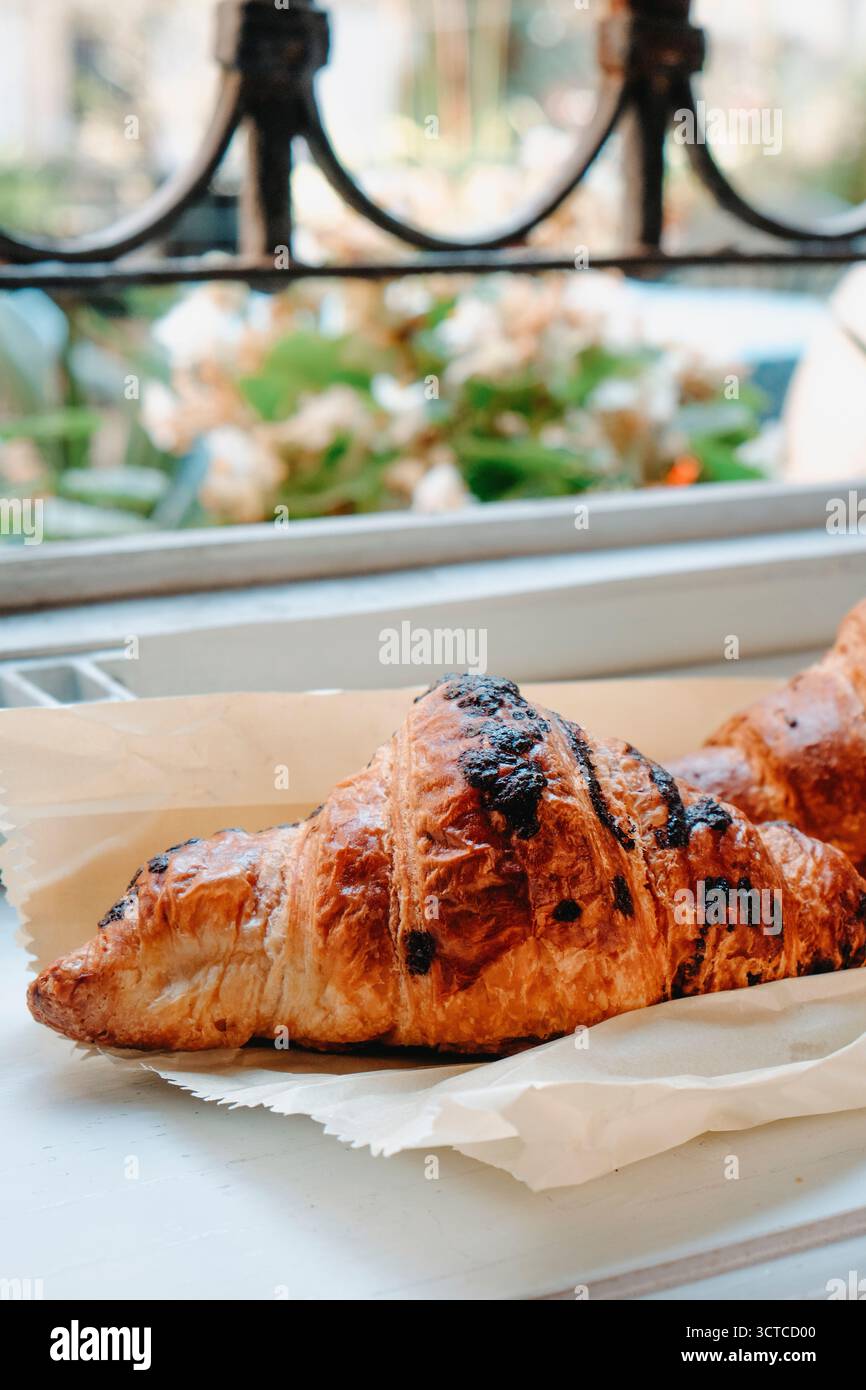 Nahaufnahme eines knusprigen Croissants mit geschmolzenen Schokoladenflecken, auf Pergamentpapier an einem Fenster platziert, mit verschwommenen Blumen und grünen Pflanzen im Hintergrund Stockfoto