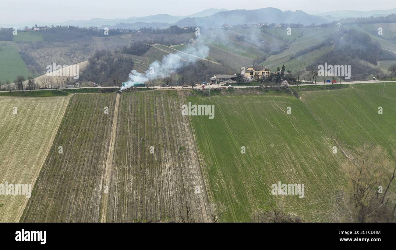 Aus der Vogelperspektive eines brennenden Traubenzweigs in den sanften Piacenza-Hügeln Italiens, umgeben von lebhaften grünen Feldern und weit entfernten Häusern, einer Rauchwolke ris Stockfoto