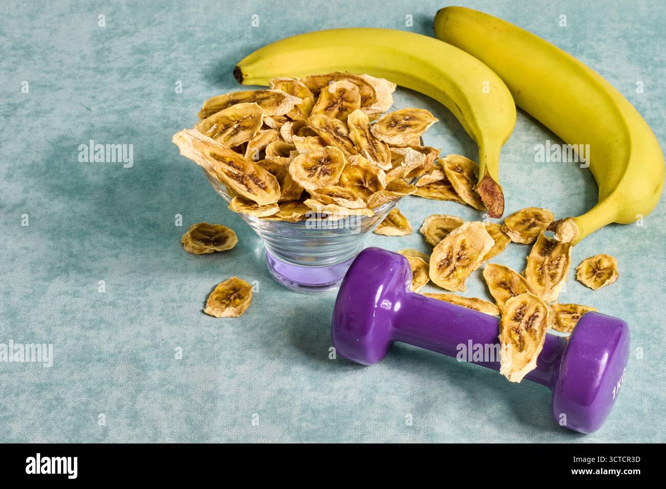 Gesundes Lifestyle-Konzept mit Bananen, Bananenchips und einer lila Hantel auf strukturiertem blauem Hintergrund, das Fitness und gesunde Snacks fördert Stockfoto