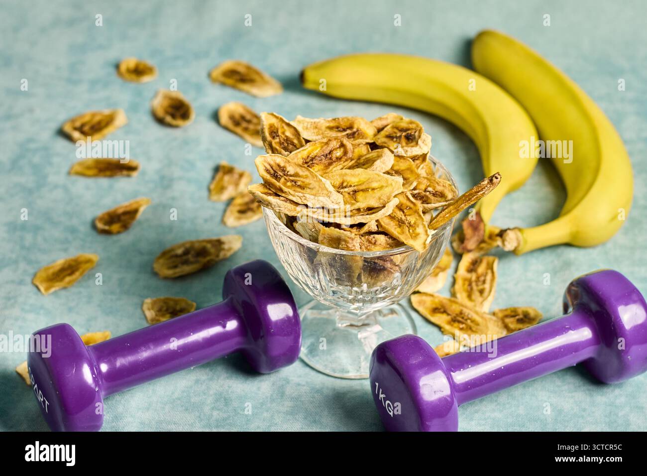 Gesundes Lifestyle-Konzept: Frische Bananen, getrocknete Bananenchips in der Schüssel und lila Kurzhanteln auf blauem Hintergrund, eine ausgewogene Ernährung für Fitnesstraining Stockfoto