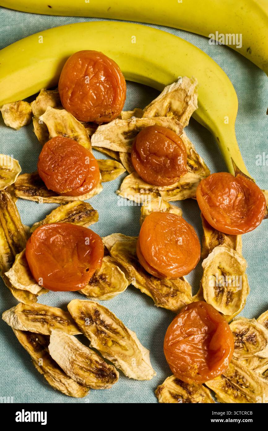 Top-Down-Ansicht von Bananenchips und getrockneten Aprikosen, die mit zwei frischen Bananen auf einer hellblauen Stoffoberfläche in einer Food-Fotografie-Umgebung gezeigt werden Stockfoto