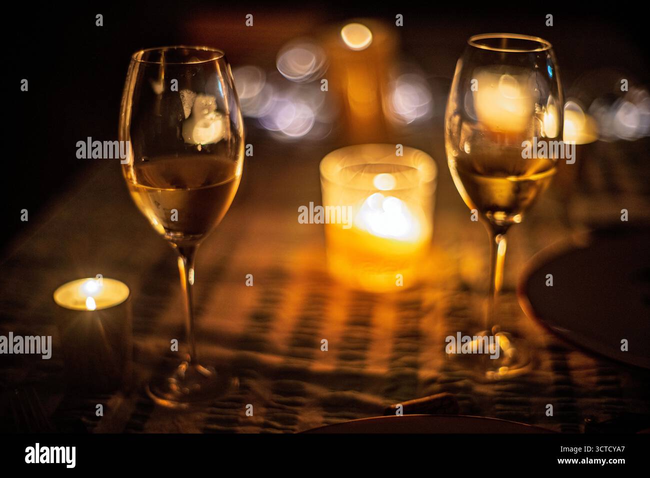 Sanftes Bokeh umgibt zwei Gläser Weißwein auf einem Tisch, der von Kerzen beleuchtet wird. Das goldene Licht verstärkt die intime Atmosphäre für einen besonderen Abend Stockfoto