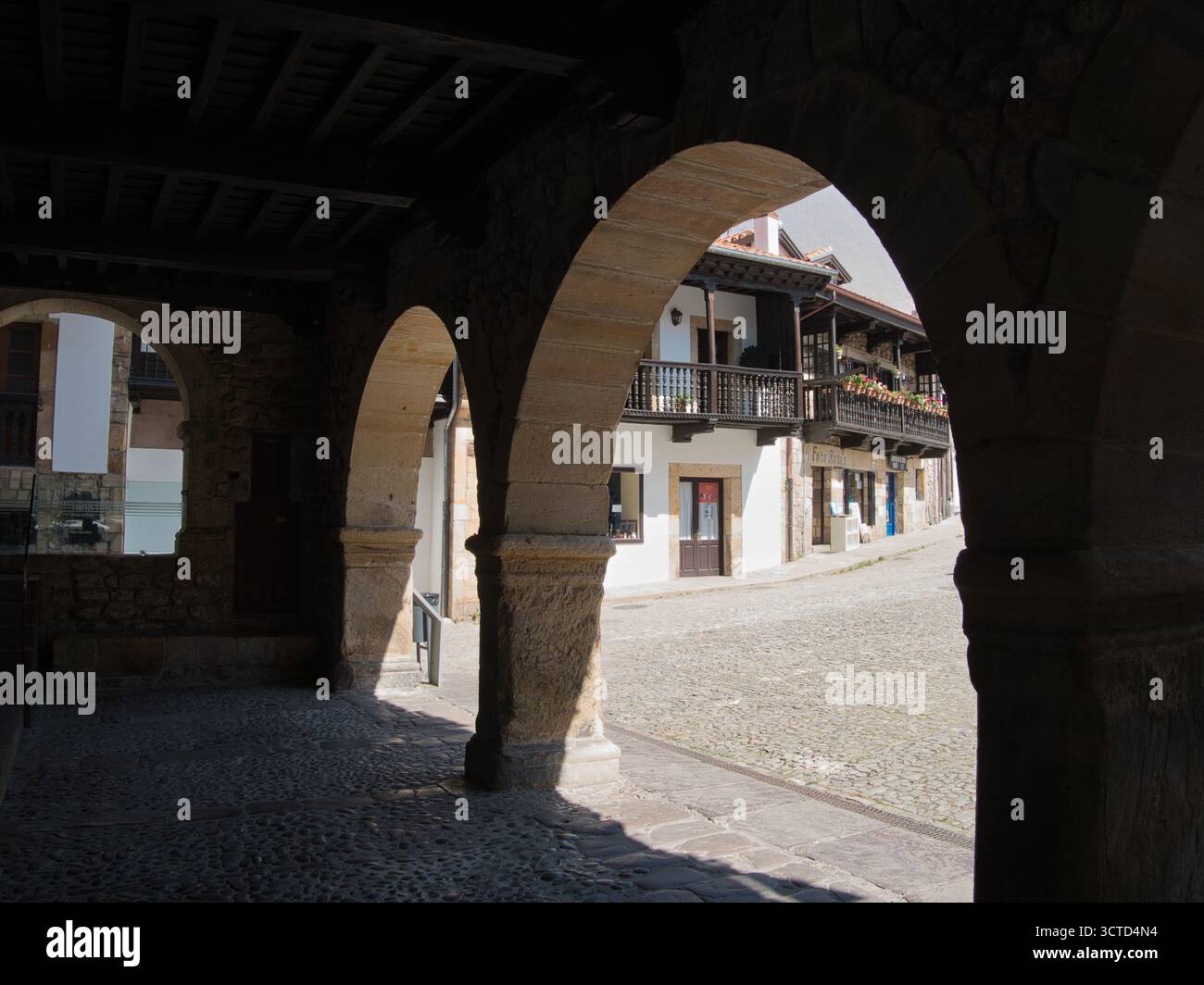 Schattige Steinbögen an der Sunny Street – Dunkle, historische Steinbögen, die einen Blick auf eine sonnenbeschienene Kopfsteinpflasterstraße und einen traditionellen spanischen Balkon bieten Stockfoto
