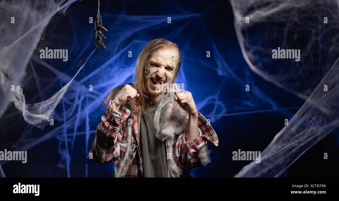 Gruseliger, gruseliger Zombie-Mann mit hässlichem Make-up, der Fäuste in aggressivem wütendem Stehen auf gruseligem Spinnennetz Hintergrund hält. Halloween-Konzept. Horrorfilm. c Stockfoto