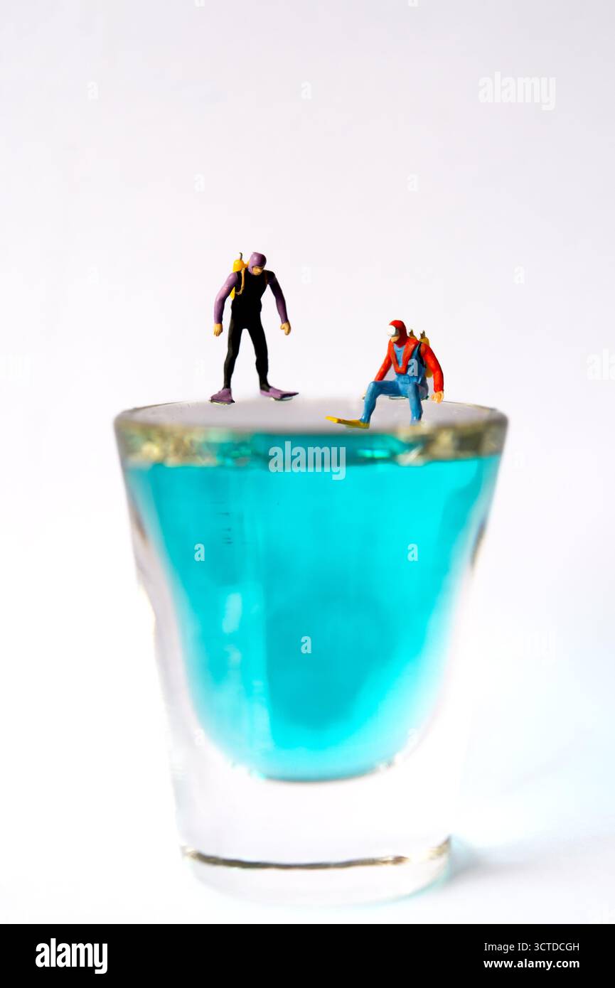 Winzige Taucher bereiten sich auf das Abenteuer vor, stehen am Rand des Glases gefüllt mit blauer Flüssigkeit – Creative Miniatur Photography Concept Stockfoto