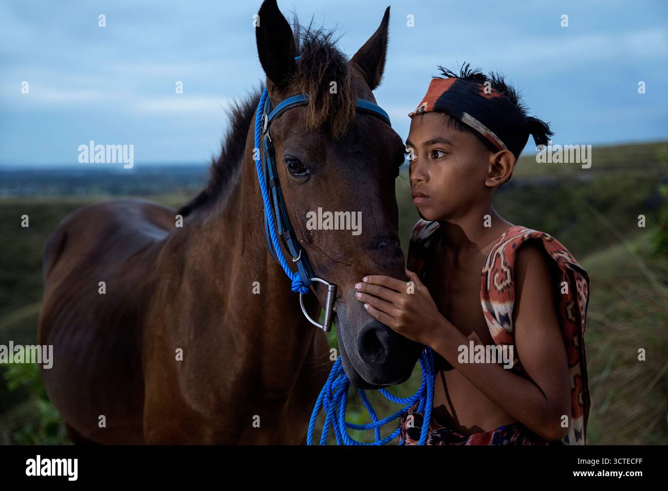 Einheimisches Kind in traditioneller Kleidung und sein Pferd auf einem grasbewachsenen Hügel im östlichen Teil der Insel Sumba in Indonesien, Junge in traditionellem Textil Stockfoto