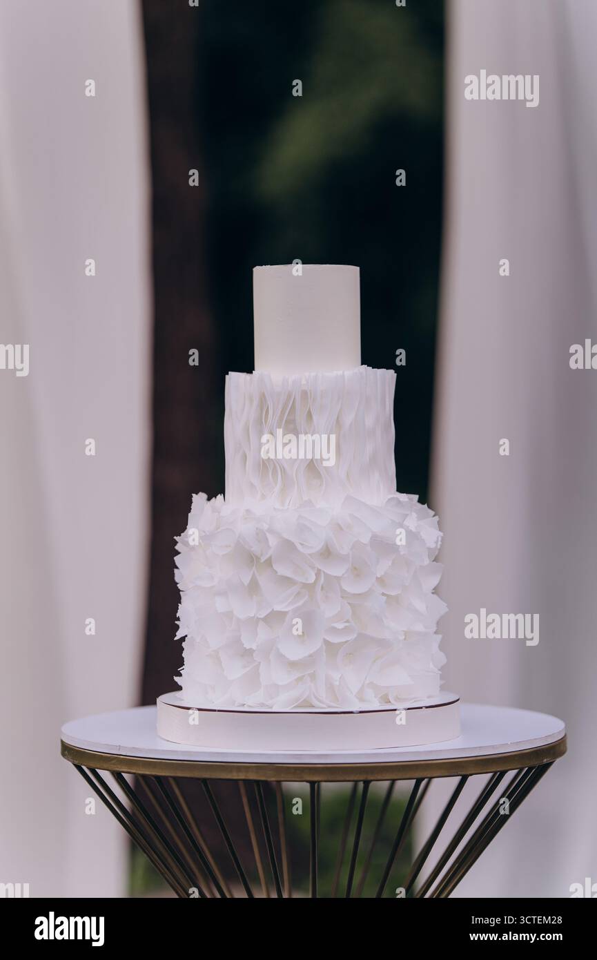 Eine atemberaubende dreistufige weiße Hochzeitstorte, elegant präsentiert auf einem stilvollen Standfuß, verziert mit zarten Rüschen und Blumendetails, perfekt für jeden Geschmack Stockfoto