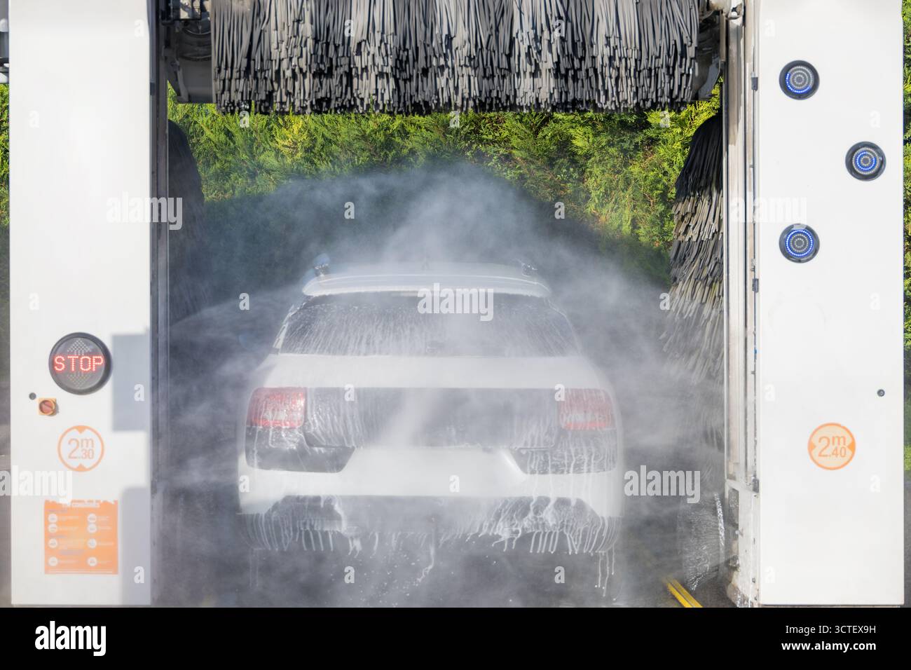 Ein mit Schaum bedecktes Auto passiert eine automatische Waschanlage mit rotierenden Bürsten und einem Wasserspray für eine professionelle Autowäsche. Stockfoto