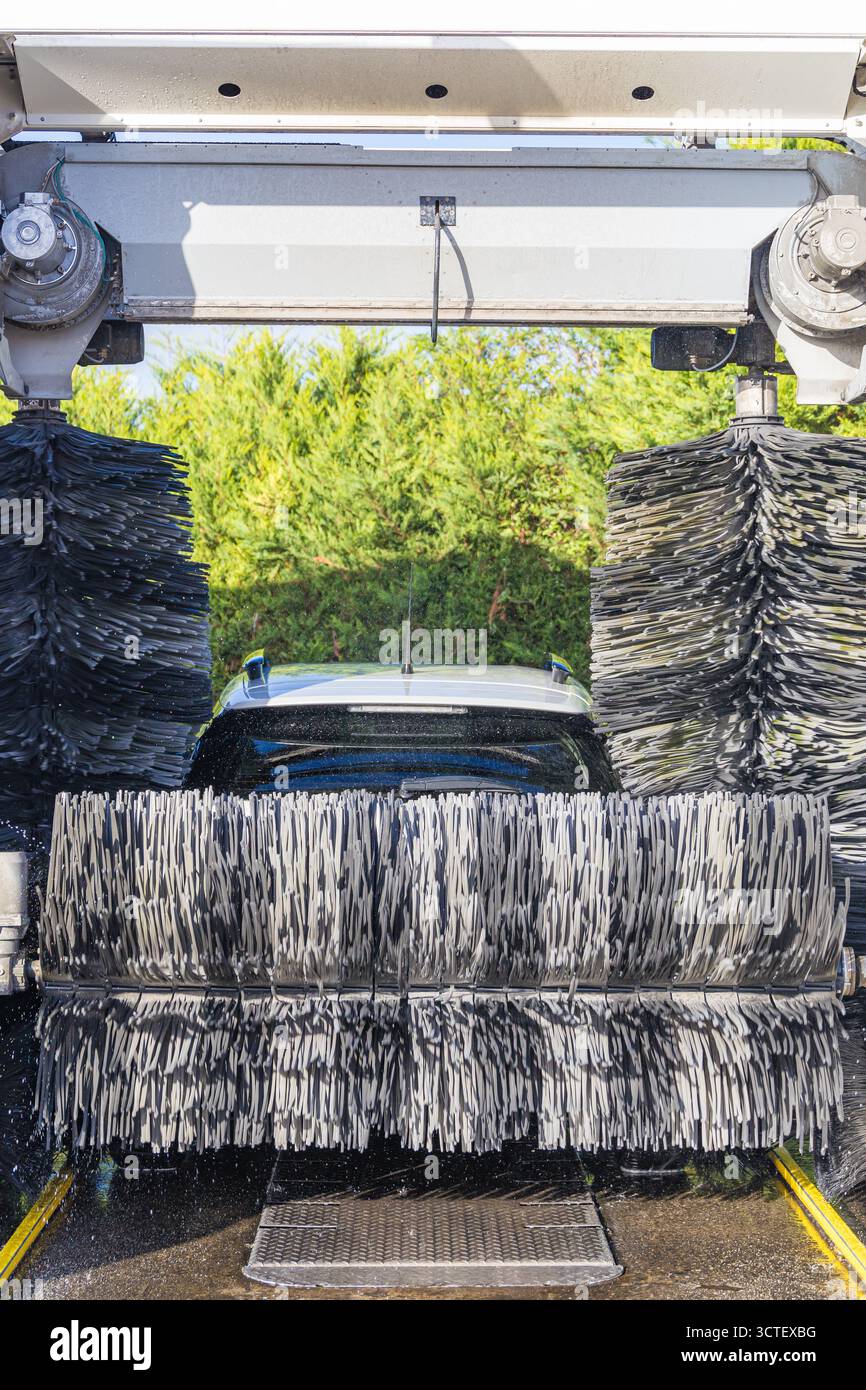 Ein mit Schaum bedecktes Auto passiert eine automatische Waschanlage mit rotierenden Bürsten und einem Wasserspray für eine professionelle Autowäsche. Stockfoto