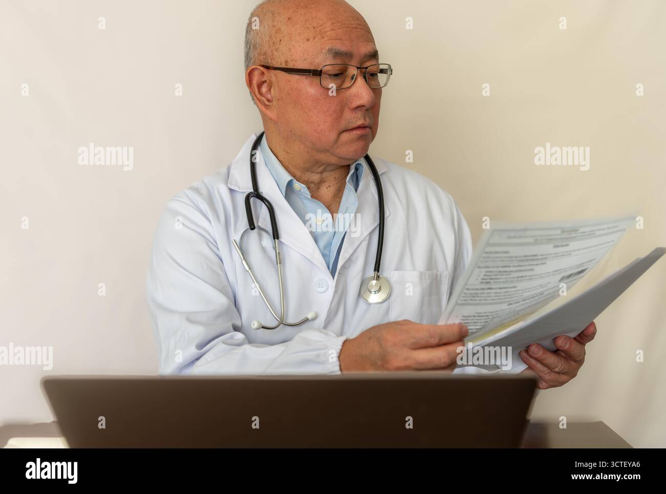 Ein Arzt mit weißem Mantel, der medizinische Dokumente liest und auf einem Laptop auf einem Schreibtisch arbeitet. Stockfoto