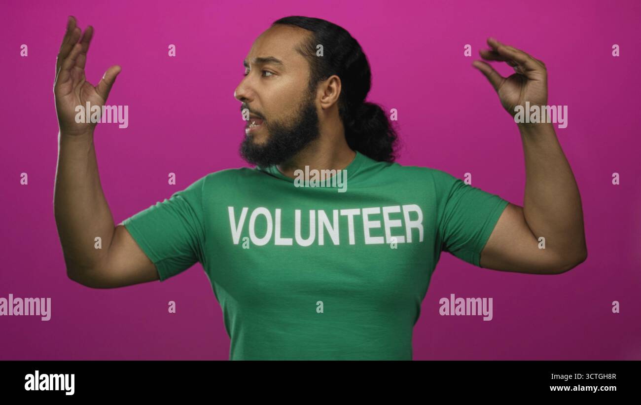 Bärtiger Mann im grünen Freiwilligen T-Shirt, das beide Arme in rosa Studio hebt; Community Outreach Enthusiasmus. Stockfoto