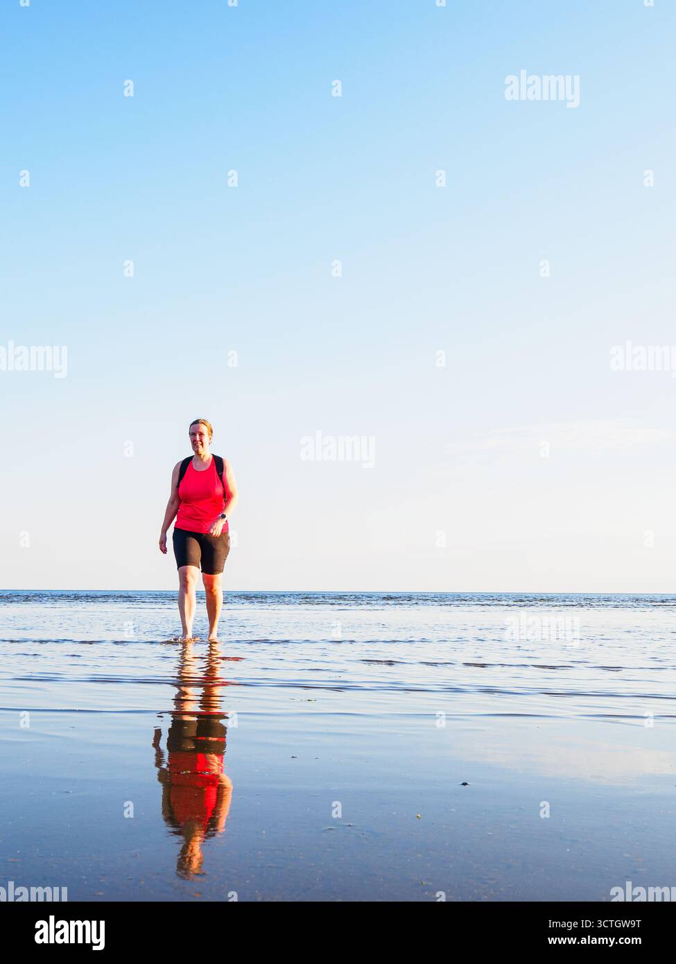 Eine Frau schlendert an einem Sommerabend an einem Strand in Schweden entlang. Das ruhige Wasser reflektiert ihre Figur, während der Himmel sich in einem sanften Blauton verfärbt und einen erzeugt Stockfoto