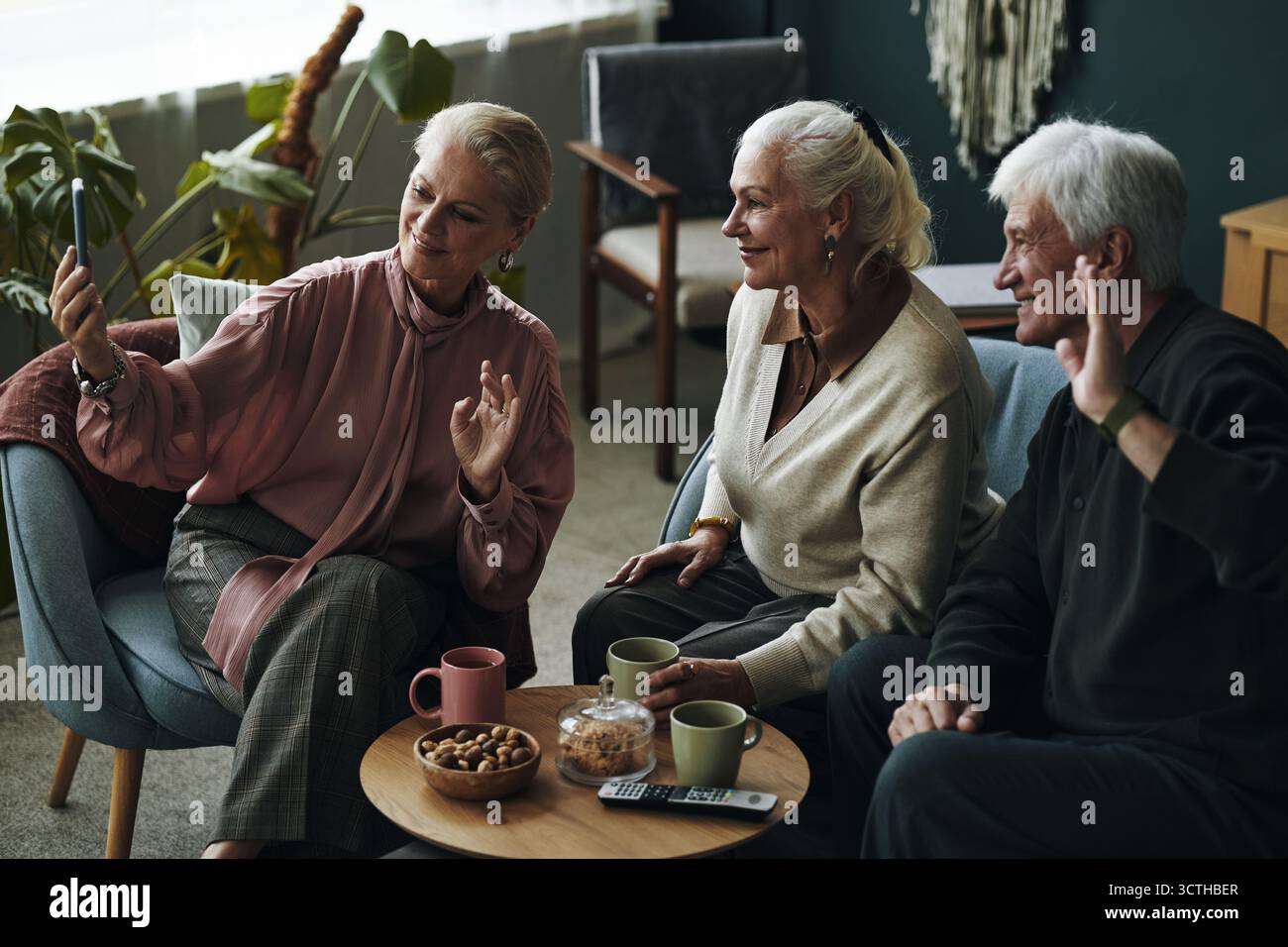 Drei ältere kaukasische Menschen sitzen zusammen und winkten während eines Videogesprächs mit Freunden zum Smartphone, zwei Frauen und ein Mann lächeln und interagieren, ein Couchtisch mit Snacks und Tassen im Vordergrund Stockfoto
