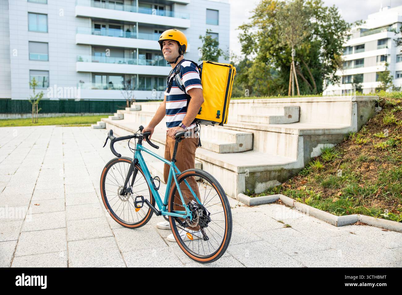 Hispanischer Mann, der einen Schutzhelm und einen gelben Rucksack trägt, neben seinem Fahrrad steht und aktiv als Kurier in einem Modus arbeitet Stockfoto