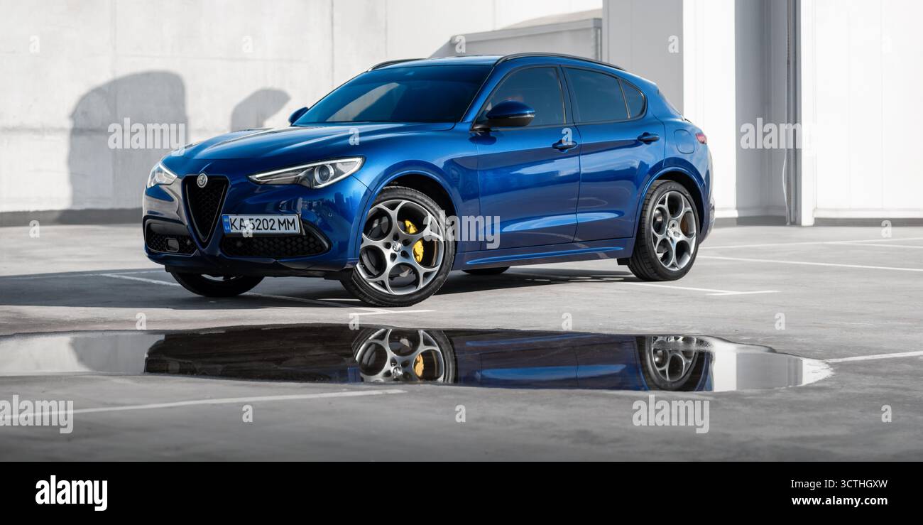 Blaues Alfa Romeo vor dem modernen Gebäude. Vorderansicht des sportlichen italienischen SUV in urbaner Umgebung, reflektiert in der Pfütze am Boden. Stockfoto