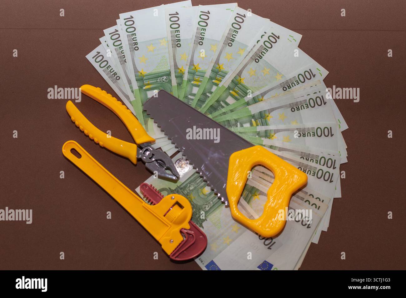 Satz gelber Spielzeugwerkzeuge, einschließlich Säge, Schraubenschlüssel und Zange, die auf einem Fächer von 100-Euro-Banknoten platziert werden. Das Konzept der finanziellen Reparatur, der wirtschaftlichen Kontrolle und der Mononie Stockfoto