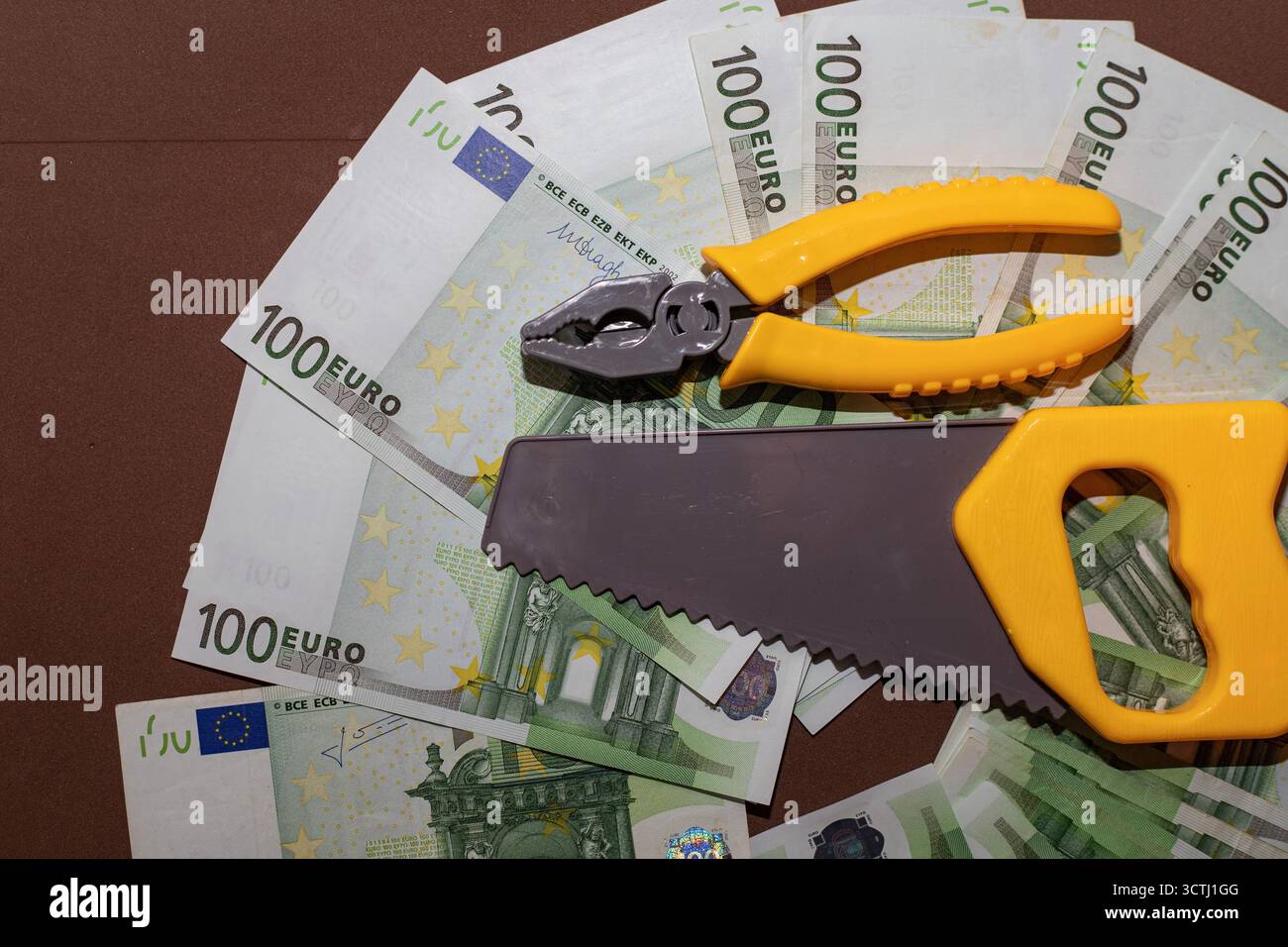 Satz gelber Spielzeugwerkzeuge, einschließlich Säge, Schraubenschlüssel und Zange, die auf einem Fächer von 100-Euro-Banknoten platziert werden. Das Konzept der finanziellen Reparatur, der wirtschaftlichen Kontrolle und der Mononie Stockfoto