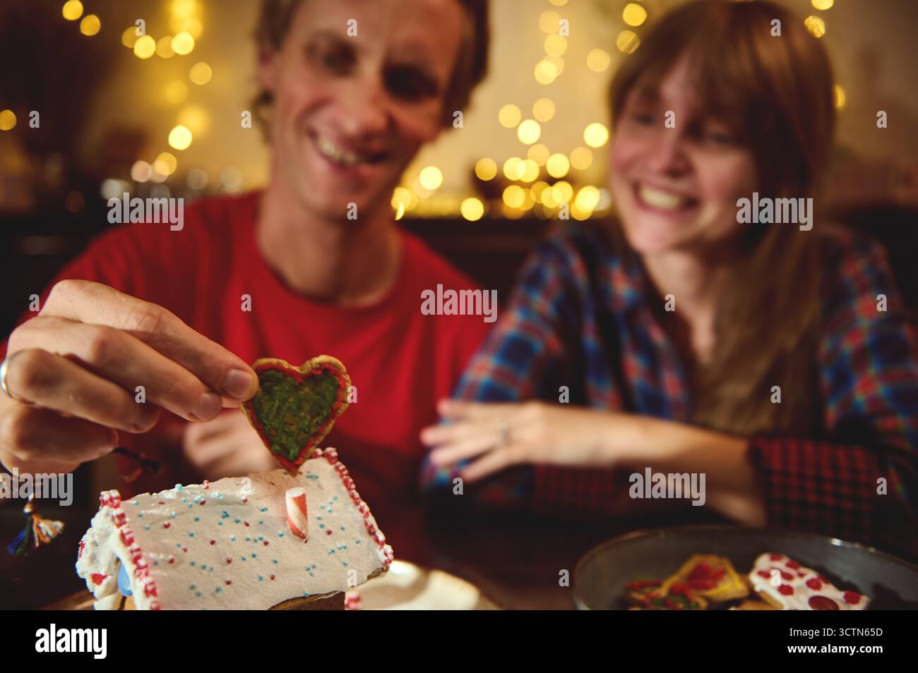 Ein lächelndes Paar genießt einen gemütlichen Urlaub, indem es einen herzförmigen Keks auf einem festlichen Lebkuchenhaus in warmen Lichtern und weihnachtlicher Atmosphäre platziert. Stockfoto Ein lächelndes Paar genießt einen gemütlichen Urlaub, indem es einen herzförmigen Keks auf einem festlichen Lebkuchenhaus in warmen Lichtern und weihnachtlicher Atmosphäre platziert. Stockfoto