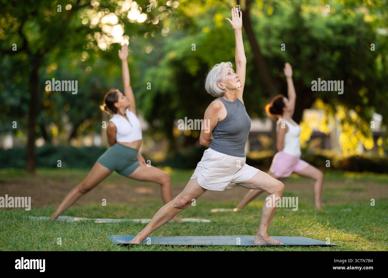 Sportliche, energiegeladene, reife Lehrerin, die Übungen auf Pilates-Matte macht und eine Variation der Kriegerposition von Yoga für Gruppenfrauen im Stadtpark demonstriert Stockfoto